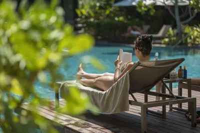 a woman sitting in a chair reading a book by a pool at Stay Wellbeing & Lifestyle Resort in Rawai Beach